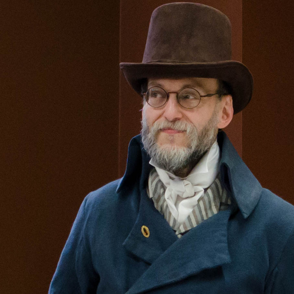 David D Levine portrait, wearing a blue Regency coat, striped waistcoat, and cravat, sporting a dashing brown top hat. 